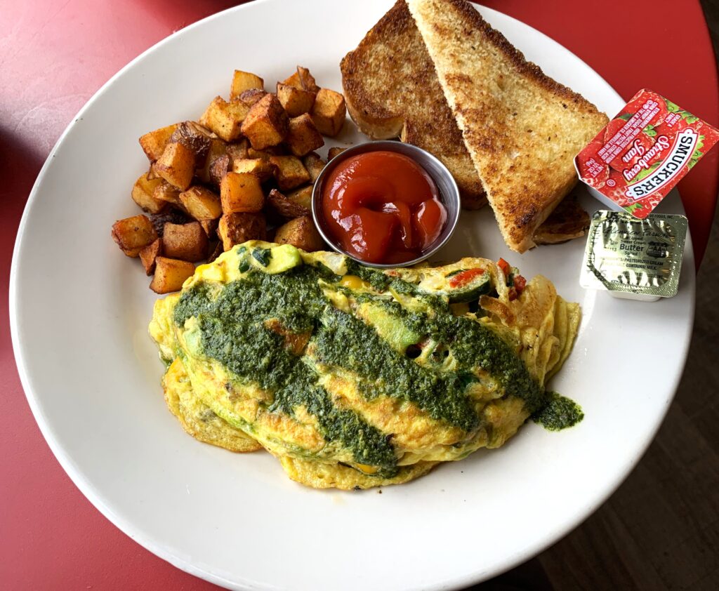 A plate with cubed fried potatoes, a vegetarian omelet topped with pesto and pieces of wheat toast, a small metal ramekin if ketchup, butter and smuckers jam.