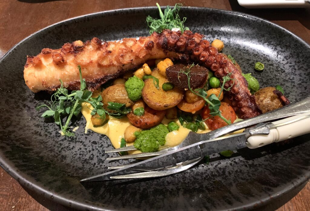 A plate with a crispy octopus tentacle, potatoes, beets, carrots, chick peas, peas and curry tahini