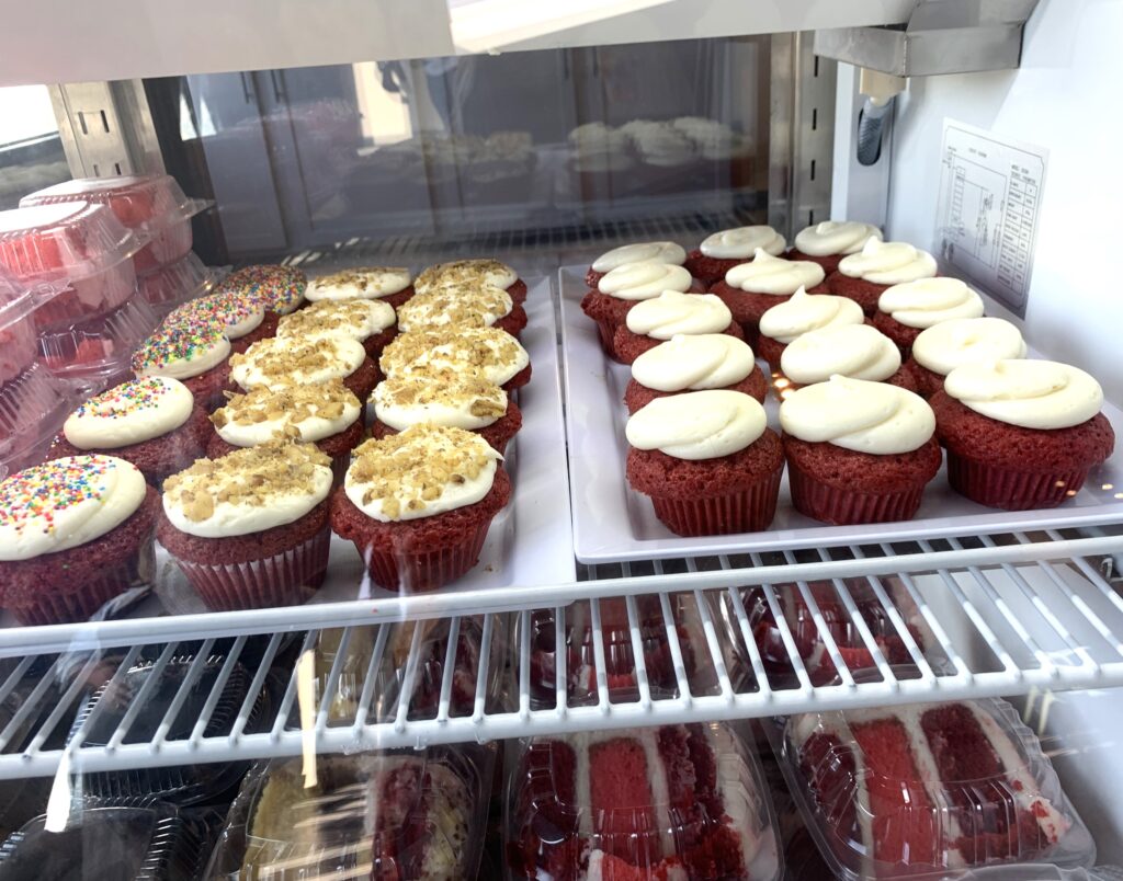Bakery store glass case with Red velvet cupcakes topped with cream cheese, another row with icing and crumbled bits and another with sprinkles, a lower shelf has slices of 3 layer cakes