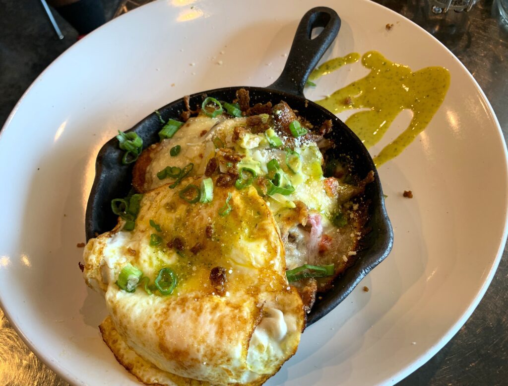 Large white plate containing a small skillet filled with layered potatoes covered in cheese and topped with scallions and fried egg, with a green aioli splash on the plate.