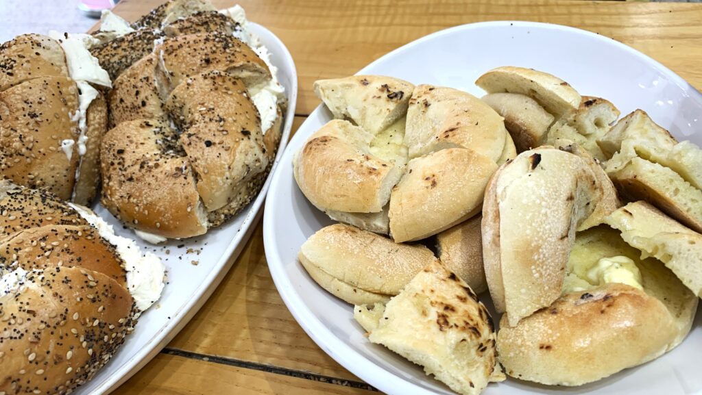 Two plates, partial plate on the left contains everything bagels sandwiched with cream cheese, with cut toasted bialys on the right sandwiched with melting butter