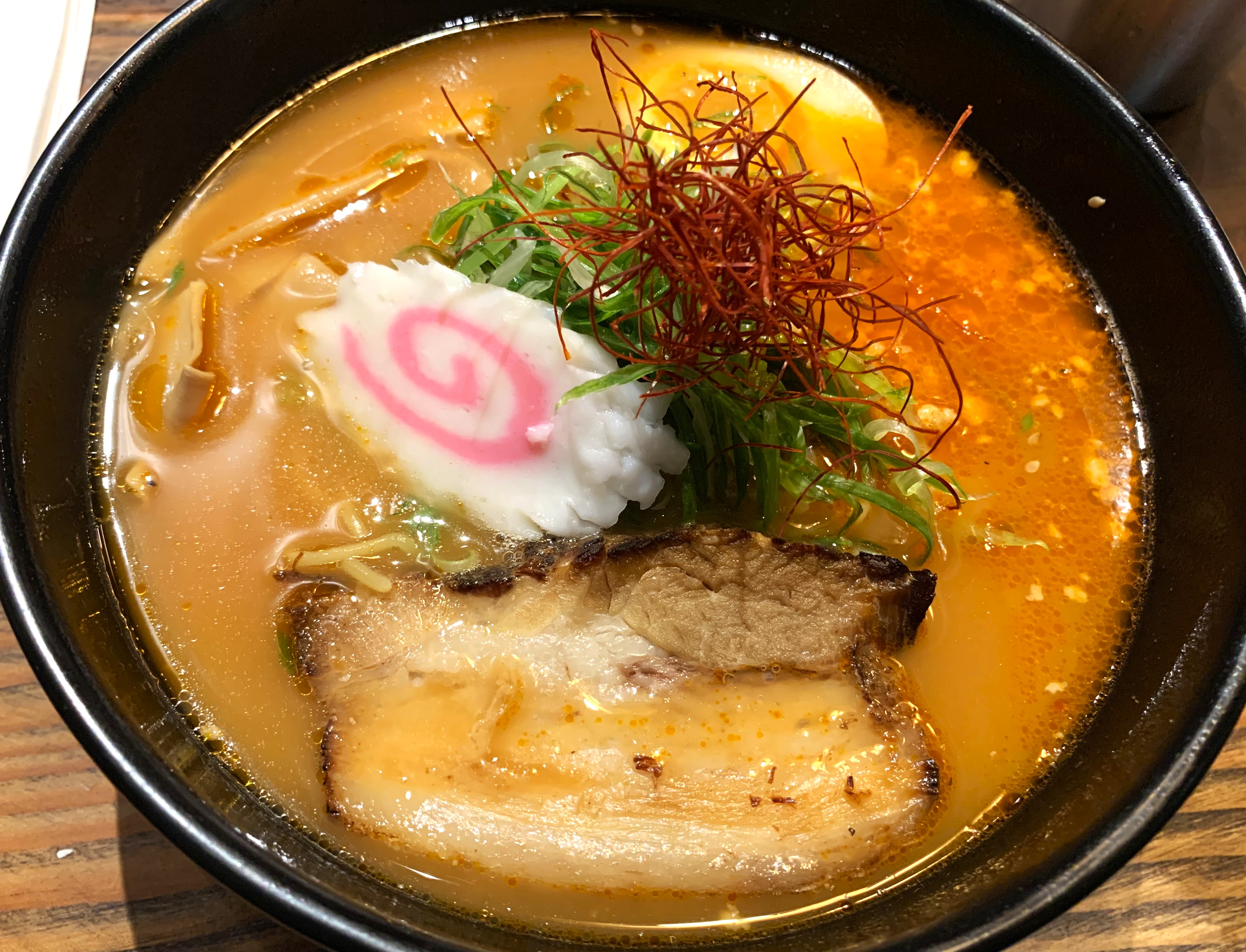 Bowl filled with a reddish broth, a piece of pork belly, a soft spiral and greens
