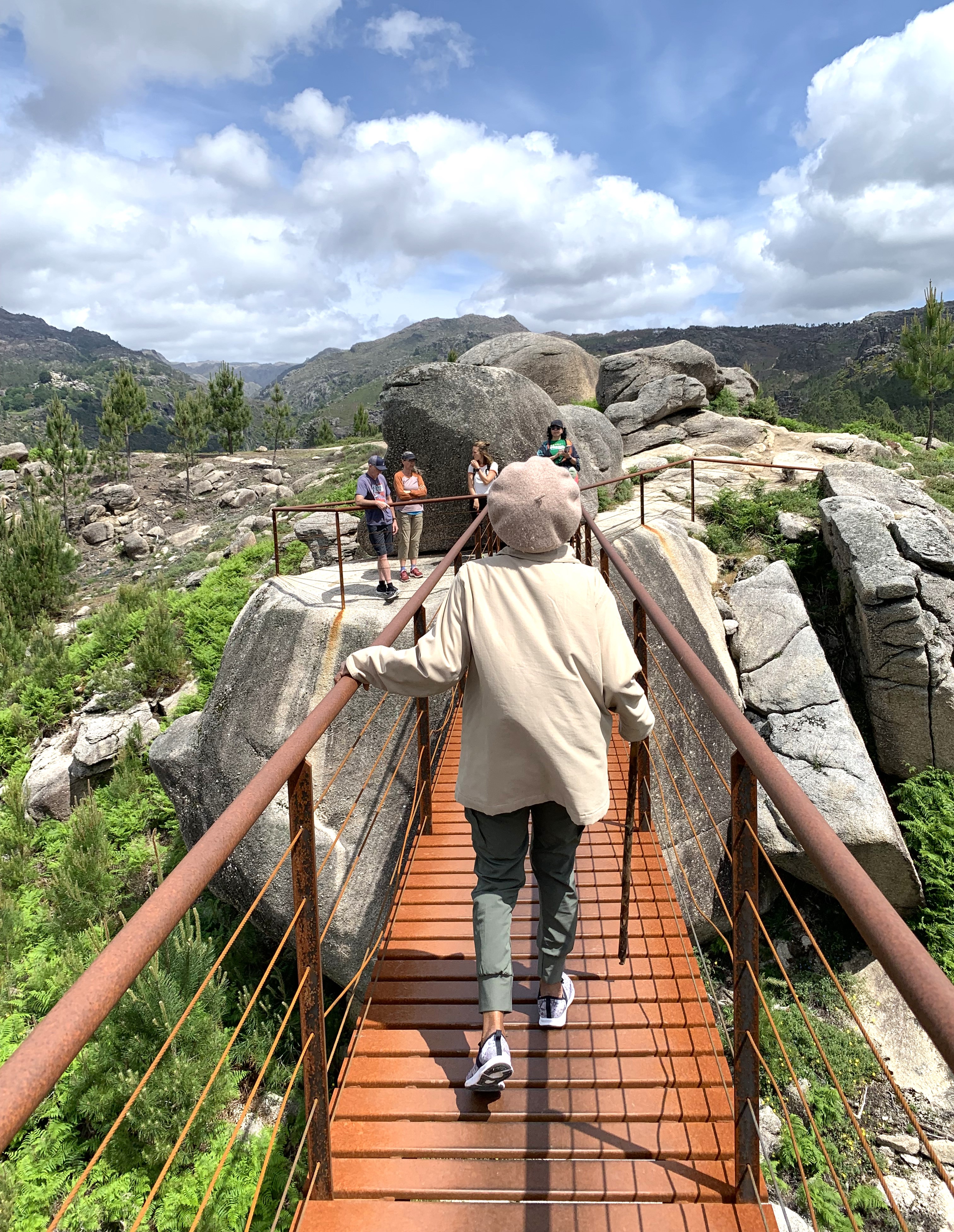 A woman with a walking stick walks across a metal bridge to a large rock look out point