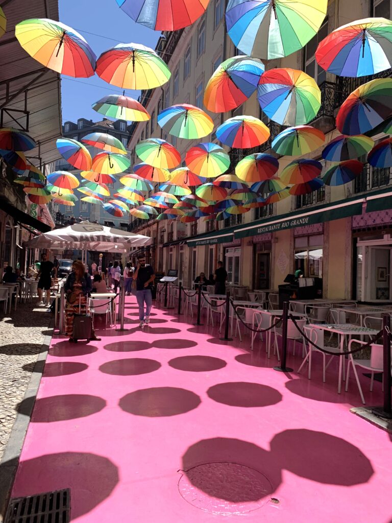 Colorful umbrellas float above a pink painted street. Tables and chairs cover the space between alongside the buildings.