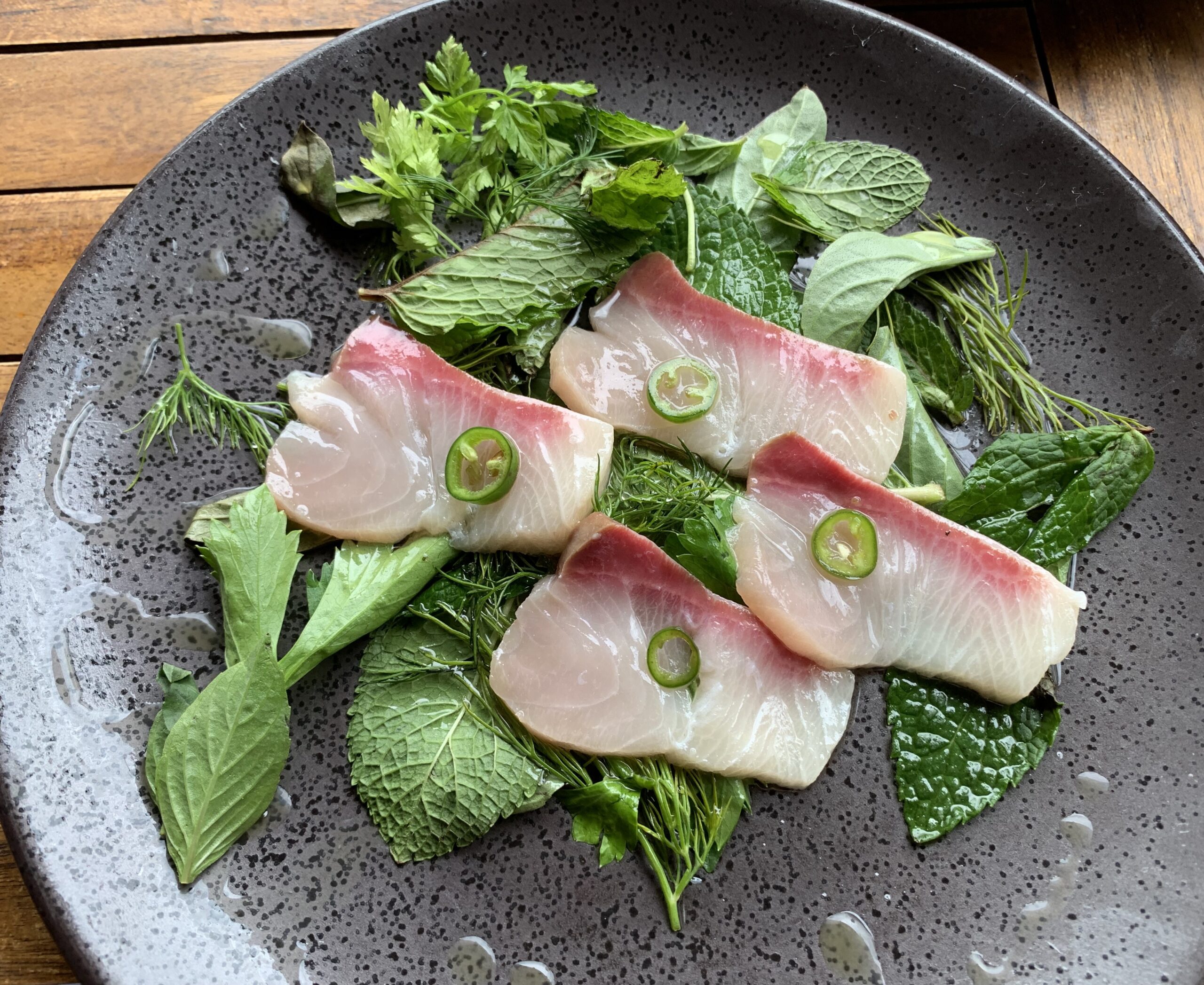 Plate with 4 sashimi sliced topped with sliced jalapeno on a bed of dill, parley and mint