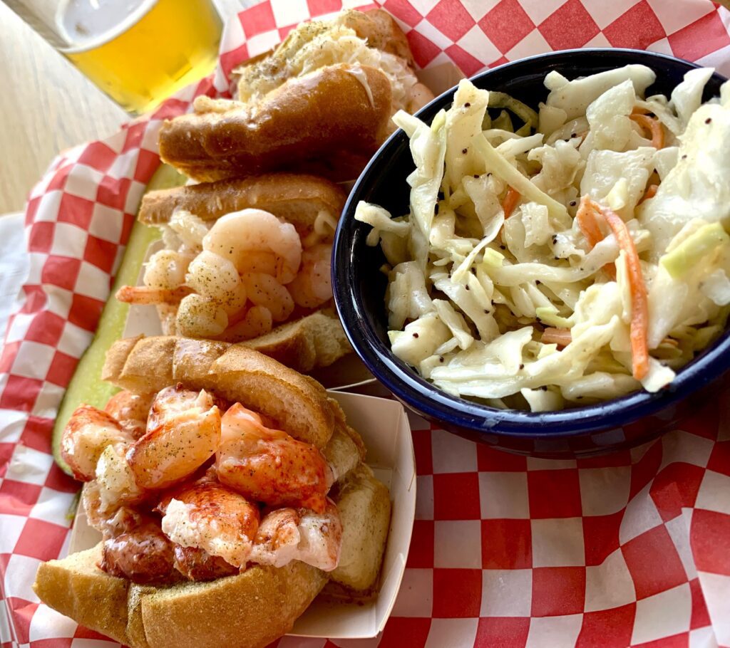 Three half sandwiches filled with lobster, shrimp and crab with a small bowl of coleslaw.