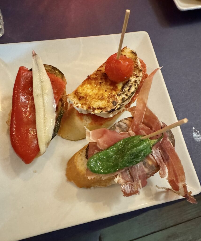 A trio of 3 pinxos on slices of a baguette, one with fish and red pepper, next with caramelized goat cheese on a tomato sauce, and last with blood sausage, ham and a green pepper