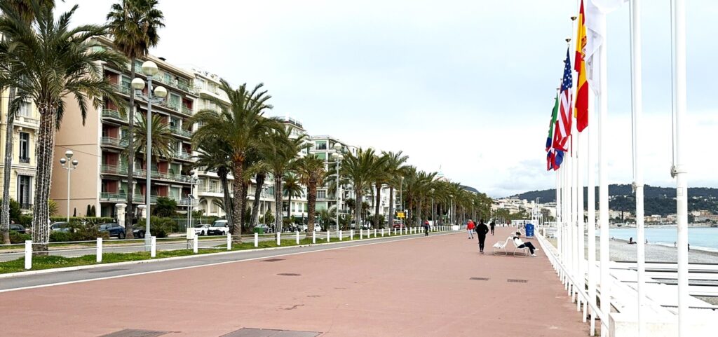 A wide boulevard follows the shoreline on the right. Flag poles line the path on the right, while a bike path edges it on the left with a road, palm trees and buildings beyond.
