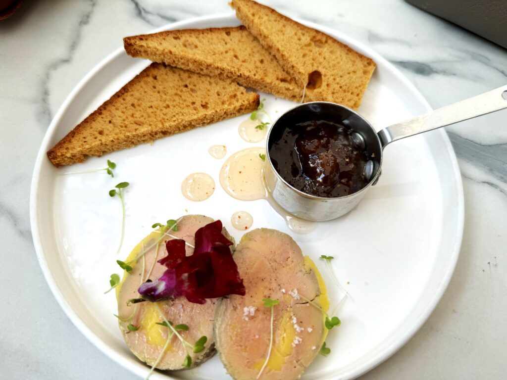 A white plate with 3 half slice pieces of ginger bread, a metal cup with fig jam and 2 slices of foie gras at the bottom