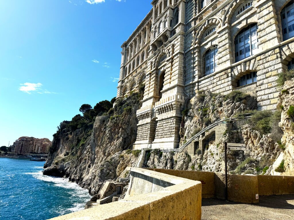 A low stone wall follows a path with blue waves beyond and stone colored building rising from the adjacent cliff, with trees further along the cliff.