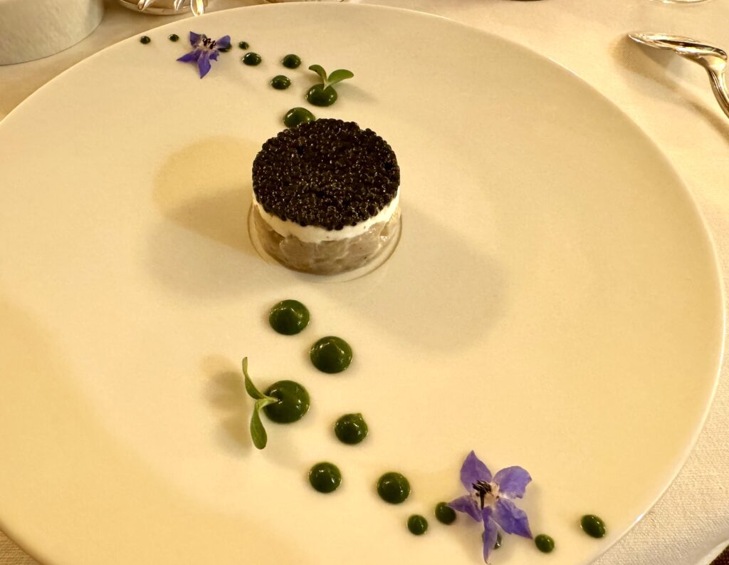 Large white plate with seabass tartar layered with oystered and topped with caviar, with green sauce dotting the plate and decorated with purple flowers.