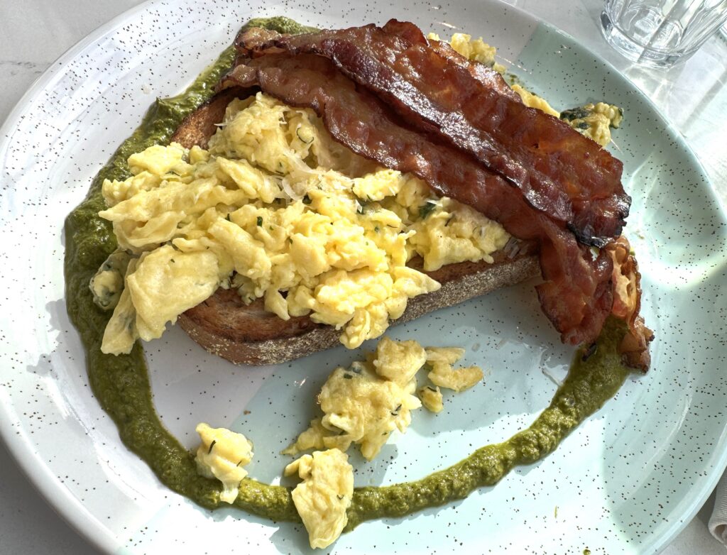 Plate with a slice of multigrain bread topped with herb scrambled eggs and cheese and rashers of bacon, within a ring of chimichurri sauce.