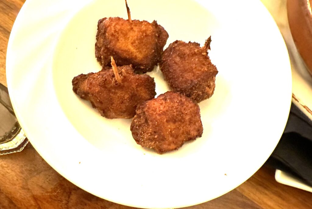 A white plate with 4 golden brown fried dates wrapped with bacon.