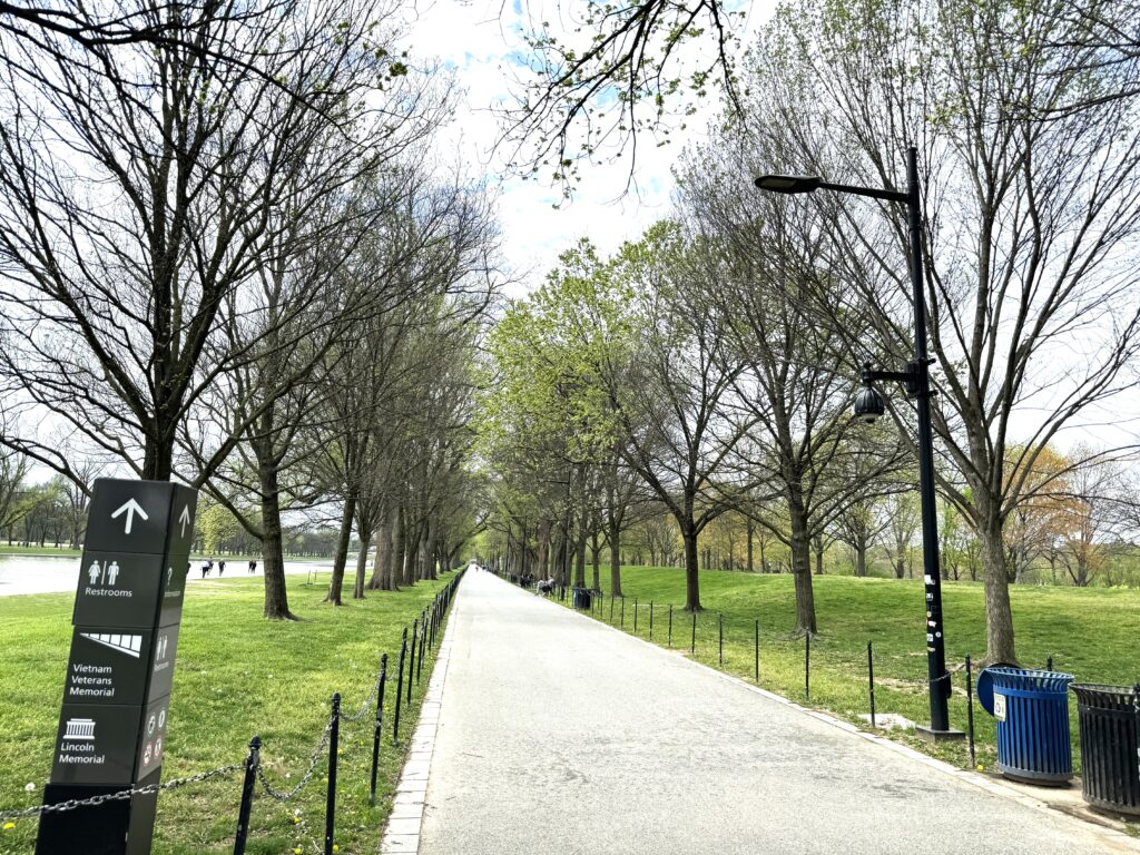 Avenue of trees parallel a paved straight path, with grass both sides of the path, and the reflecting pool on the left. A marker on the left shows directions to restrooms, the Vietnam Veterans Memorial and the Lincoln Memorial.