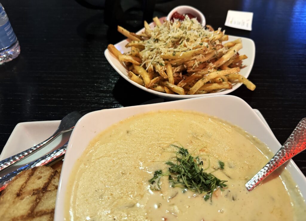 Partial bowl of Lobster and corn chowder topped with green herbs, and a plate of shoestring french fries topped with herbs and parmesan cheese.