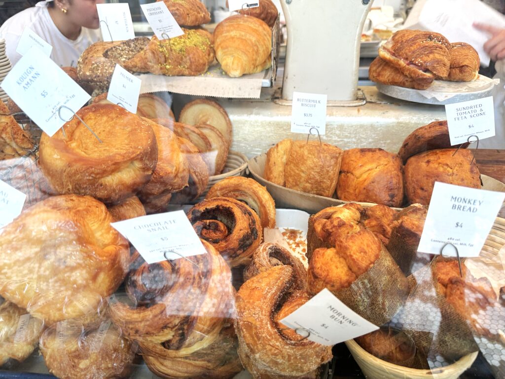 Display case filled with pastries in baskets, bowls and platforms including monley bread, Kouign amann, chocolate snail, sundried tomato & feta scone, morning bun, buttermilk biscuit and pistachio croissant.
