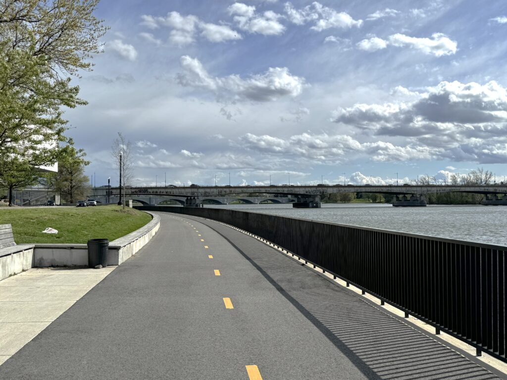 Paved running/bike trail divided by a yellow line runs along and curves to follow the river on the right. A stone arched bridged lies beyond. A black metal fence divides the trail and the river.